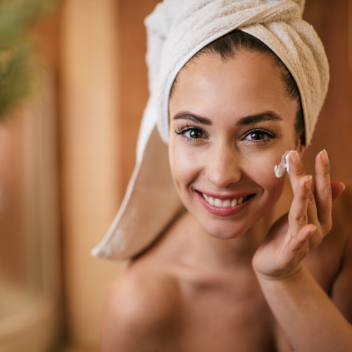 Femme souriante appliquant crème visage, serviette sur tête.